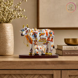 Decorative cow figurine with colorful designs on a wooden surface, featuring the brand 'Kraphy'. thumbnail